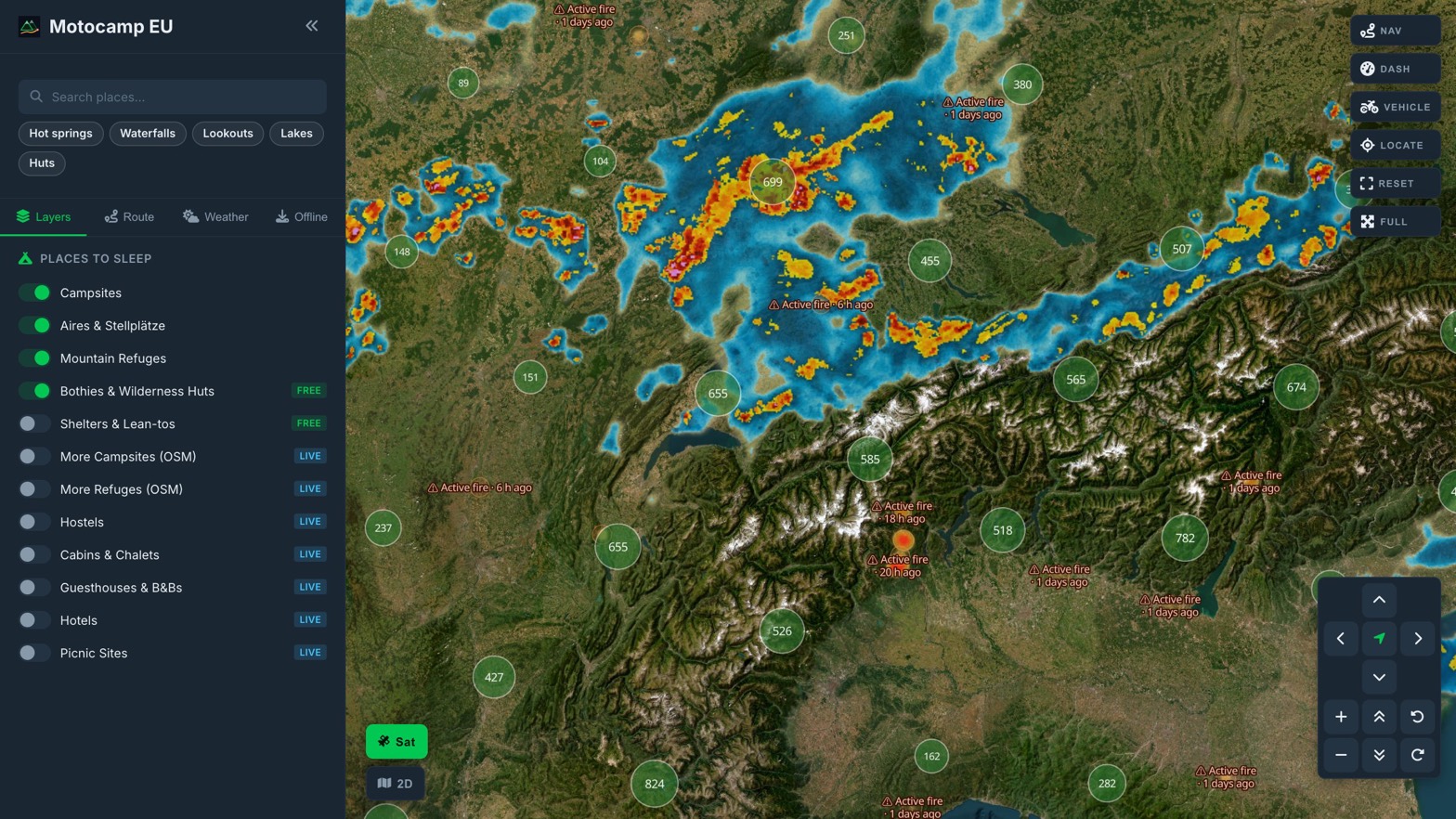 Live rain radar over the Alps in Motocamp EU: a band of blue, yellow and red cells from the Swiss plateau across Bavaria and Austria, drawn over the satellite basemap with campsite clusters and fire labels still readable on top.